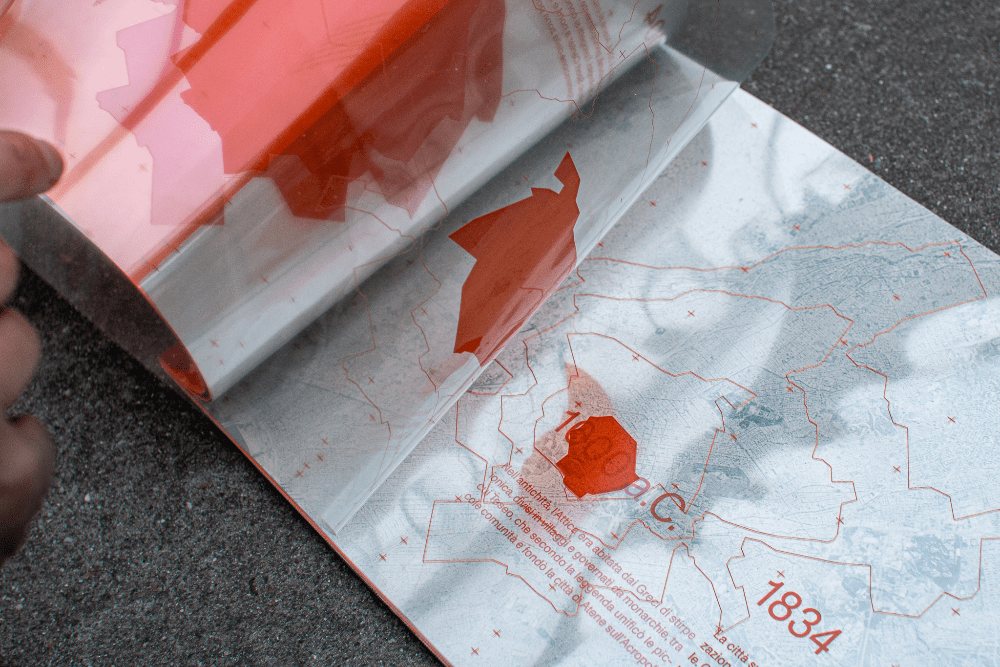 Close-up of the Anti-Atene publication being opened, a translucent red acetate sheet folding back to reveal a historical map of Athens with orange geographic outlines