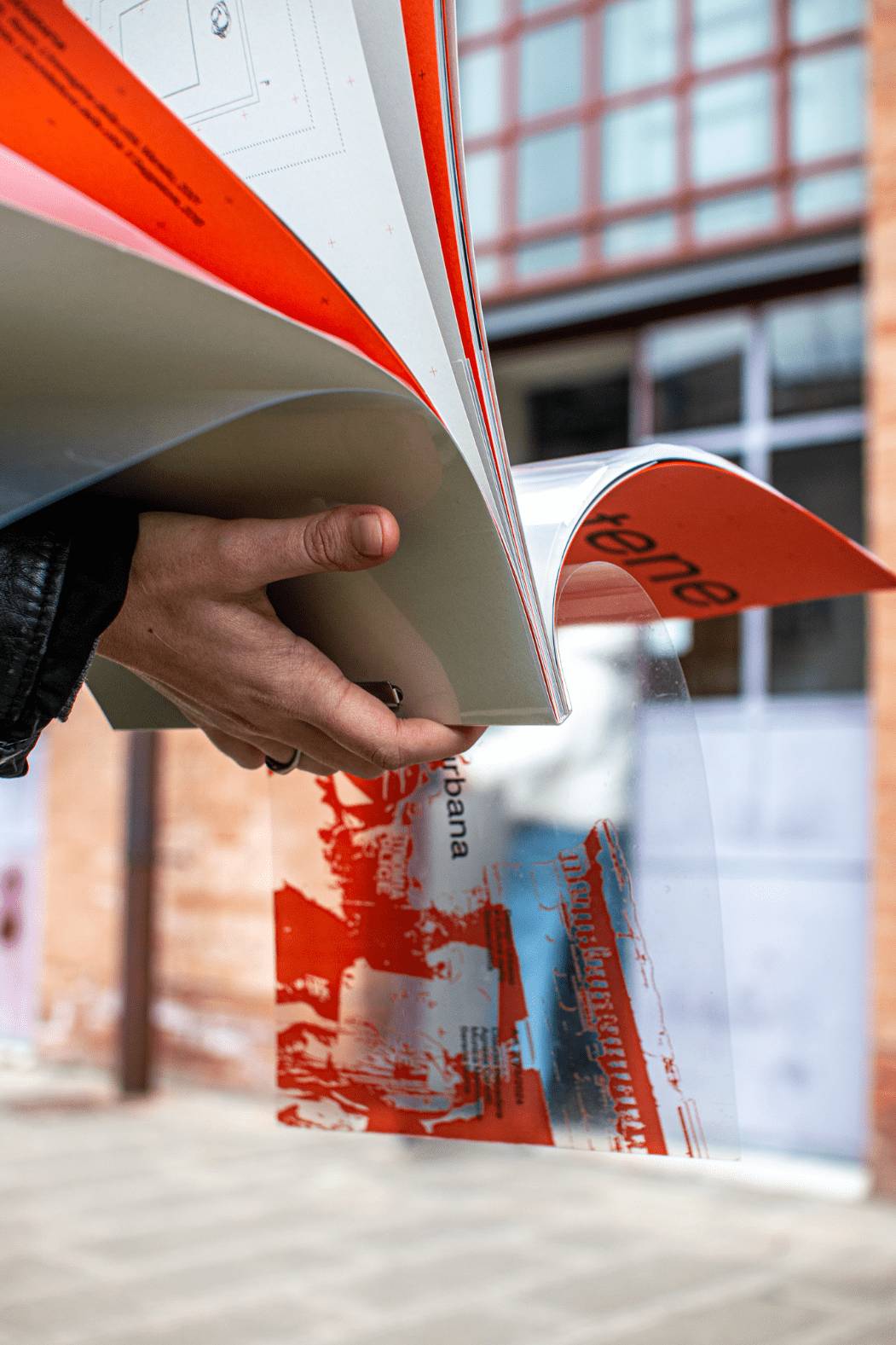 Hand holding open the Anti-Atene publication, pages fanning outward, red and gray editorial spreads visible against an urban background